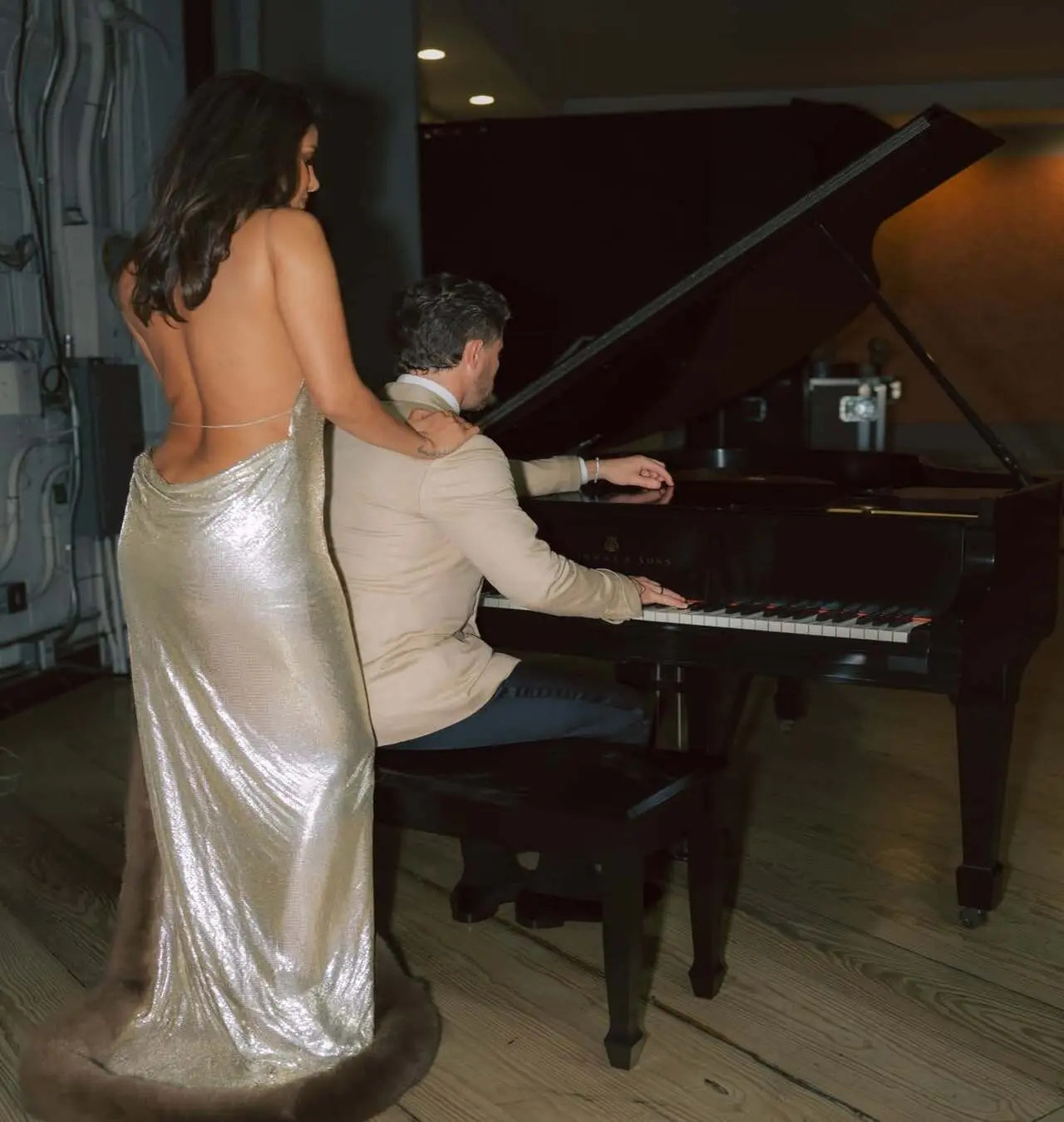 Priyanka shows off the backless detail of her gown and she olaces her hand on Nick's shoulder, while he serenades her by playing the piano